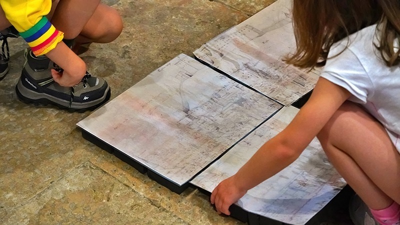 Descrição da imagem: nesta fotografia vemos de costas e de cócoras, uma criança a montar as várias "peças" do puzzle que compõem a imagem de Leiden, espalhadas no chão de pedra da Sala Ogival.