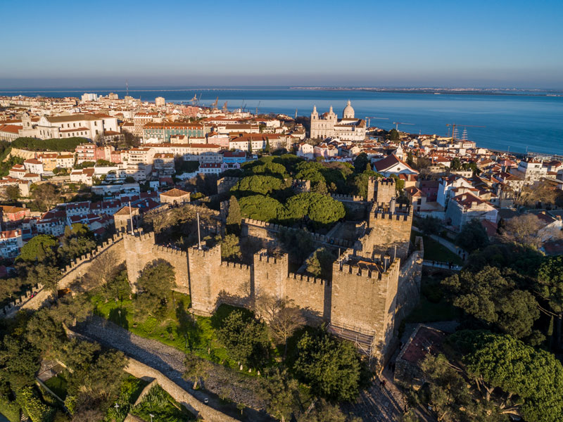 Descrição da imagem: fotografia tirada com drone do Castelo de São Jorge, onde vemos em primeiro plano o castelo, em segundo plano o casario de Lisboa, com destaque para o Mosteiro de São Vicente de Fora, e ao fundo, o Mar da Palha e o céu azul. ©Sergiy Scheblykin