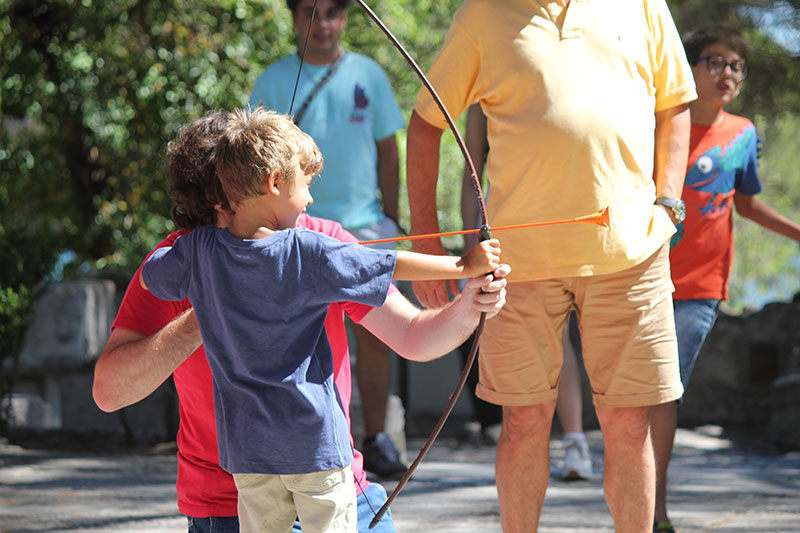 Descrição da imagem: fotografia de um menino a segurar um arco e flecha com a ajuda do técnico de serviço educativo que acompanha a atividade.