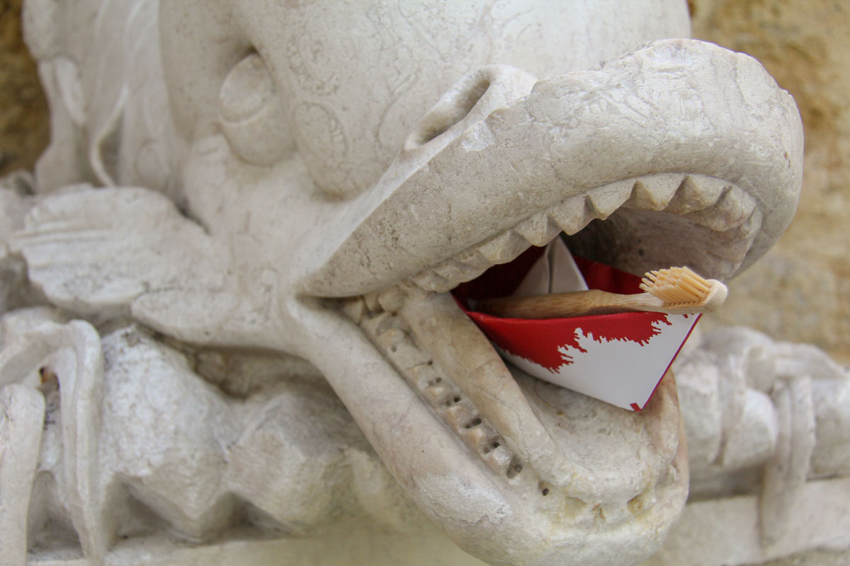 Descrição da imagem: fotografia de uma estátua de um animal marinho, de boca aberta com os dentes à mostra, e dentro da boca uma escova de dentes e um barquinho de papel.