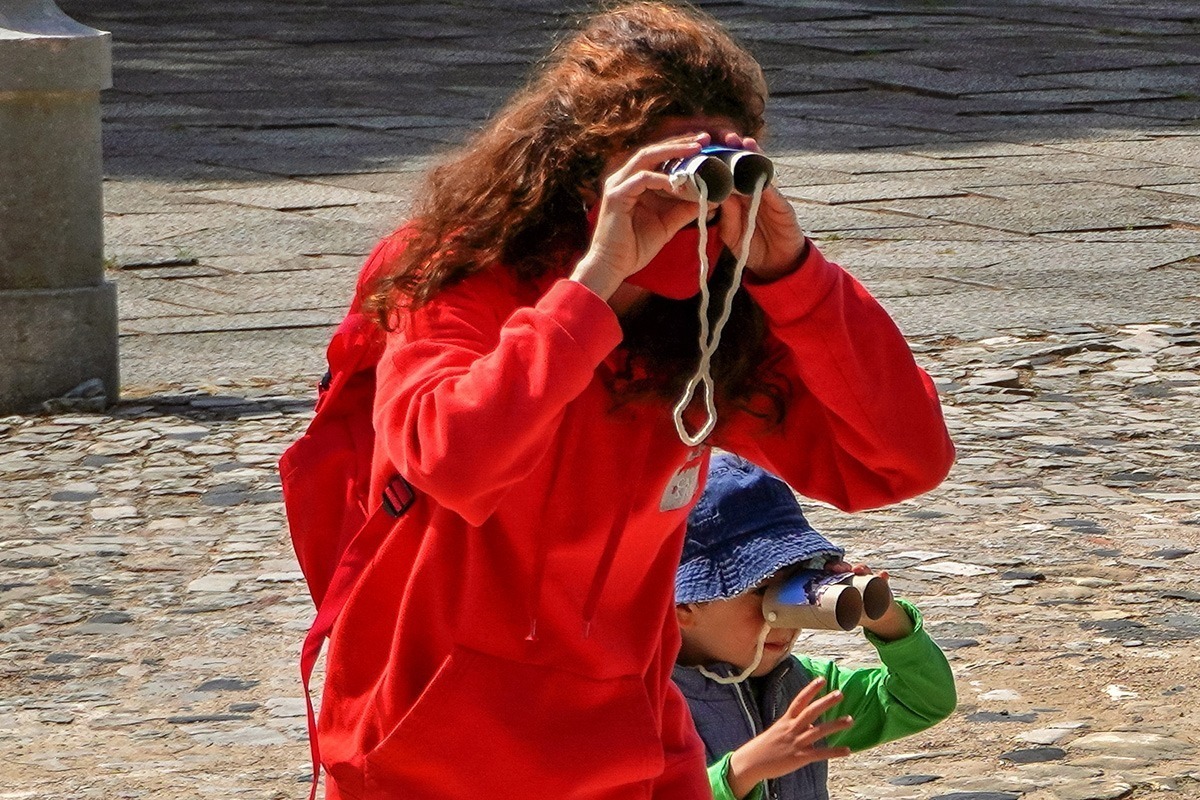Descrição da imagem: fotografia de uma técnica do serviço educativo do castelo, acompanhada por um dos miúdos participante na oficina, ambos na Praça de Armas do Castelo a observar com uns binóculos feitos de rolos de papel higiénico.